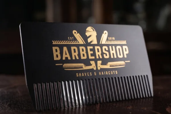 A black metal barbershop business card die-cut into a comb, with a gold foil logo.