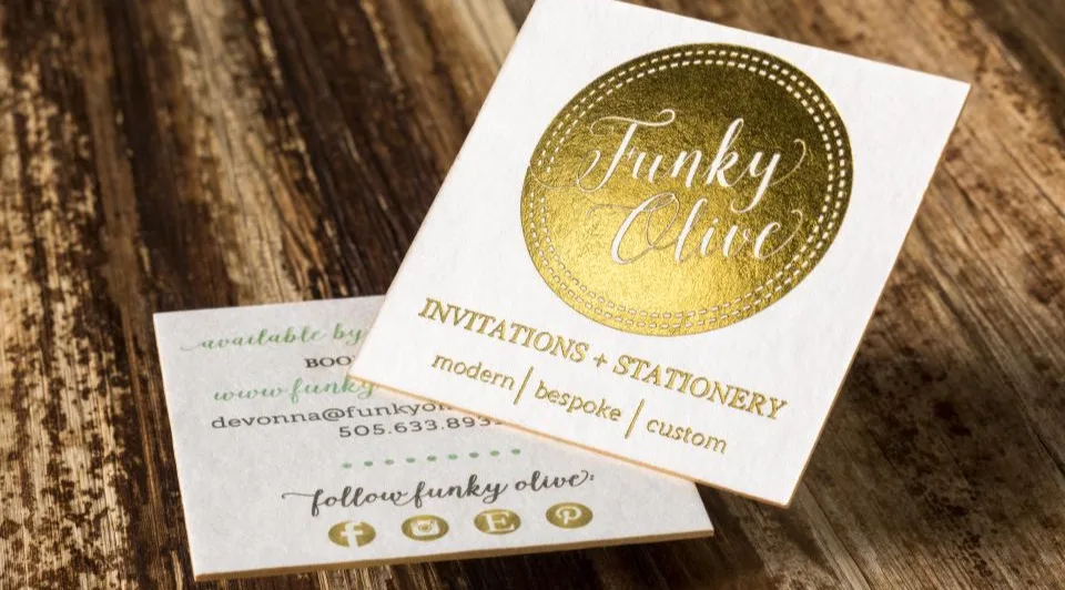 Cream cotton wedding stationery card with a gold foil circular "Funky Olive" emblem on a wood surface.