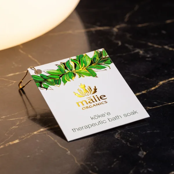 A smooth cream hang tag with a green foil botanical design, clipped to a gold safety pin.