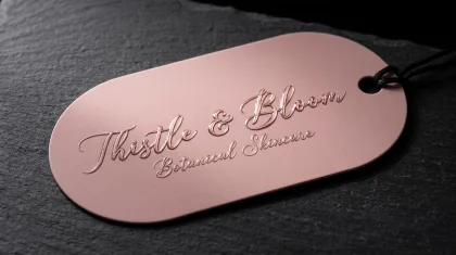 A rose-gold hang tag with glossy raised lettering reading 'Thistle & Bloom Botanical Skincare' on a dark slate surface, corded through a punched hole.